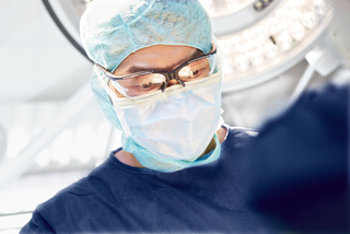 Surgeon in mask and cap focused on procedure under bright surgical lights.