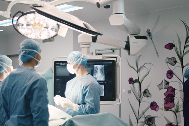 Surgeons in a modern operating room with digital screens and floral wall art, working around a patient.