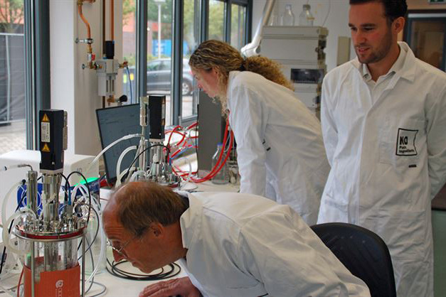 Three scientists in lab coats work with laboratory equipment; one leans in closely, while two others observe.