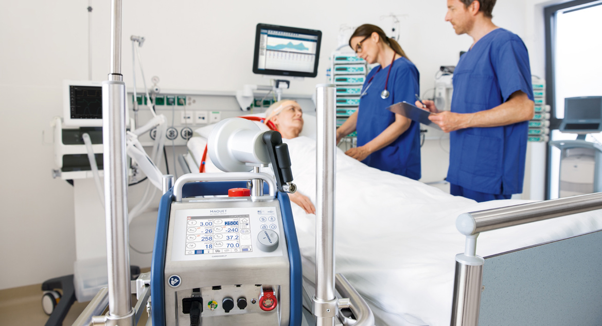 Medical staff in blue scrubs attend to a patient in a hospital bed, with medical equipment in the foreground.
