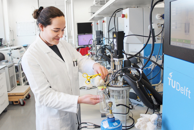 A scientist in a lab coat works with equipment in a laboratory, using a glass container and yellow clamp.