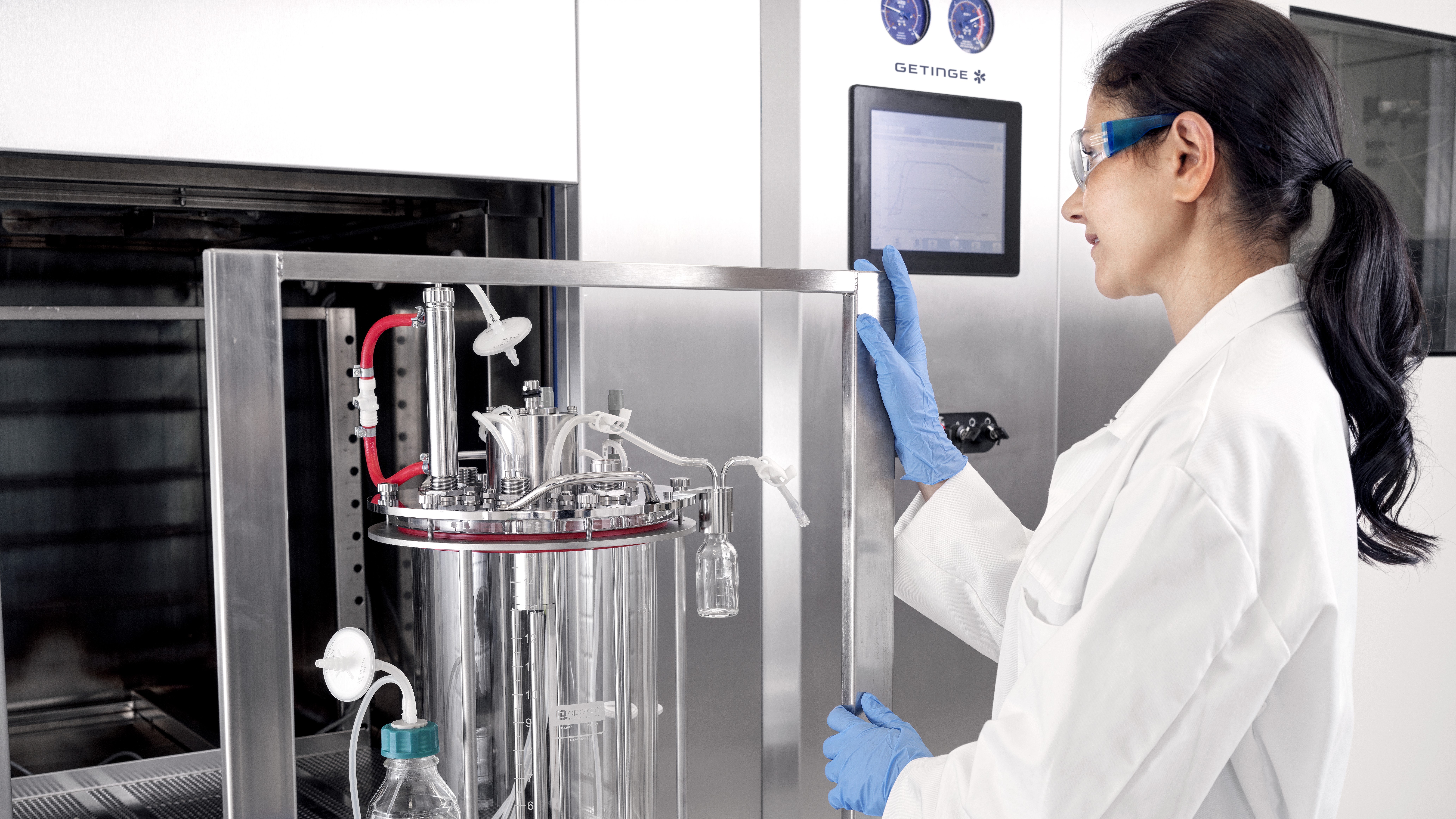 A scientist in a lab coat and safety glasses operates a stainless steel lab machine with a digital display.