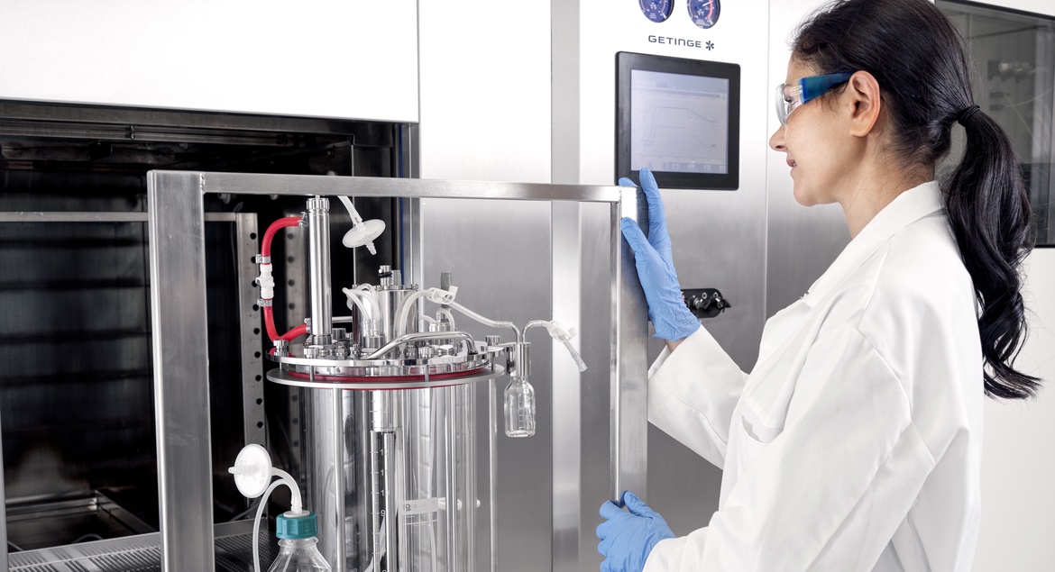 A scientist in a lab coat and safety glasses operates a stainless steel lab machine with a digital display.