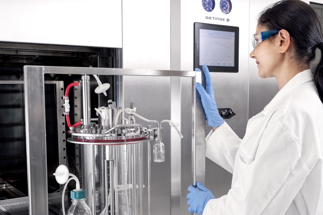 A scientist in a lab coat and safety glasses operates a stainless steel lab machine with a digital display.