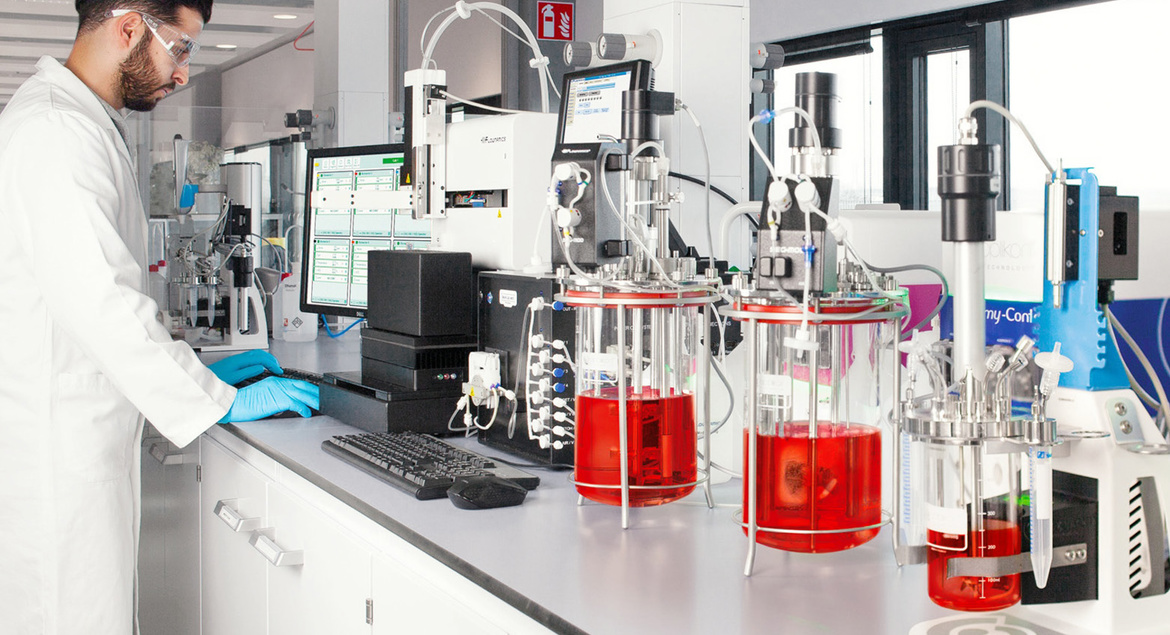 A scientist in a lab coat works at a computer beside equipment with red liquid in a modern laboratory.