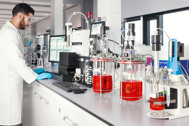 A scientist in a lab coat works at a computer beside equipment with red liquid in a modern laboratory.