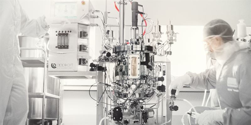 Two scientists in protective clothing operate complex lab equipment in a sterile laboratory environment.