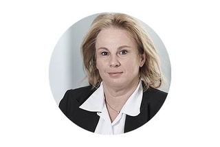 En person med blont hår i kostym, mot en ljus bakgrund.