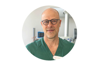 En person som bär glasögon och gröna skrubbar står i en medicinsk miljö och ler mot kameran.