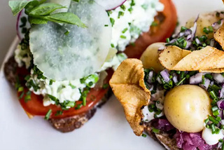 Två smörgåsar med öppen ansikte med olika pålägg inklusive potatis, tomater, grönt och chips på en vit tallrik.