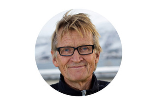 Leende person med glasögon och kort hår, klädd i en mörk jacka, utomhus med en snöig bakgrund.