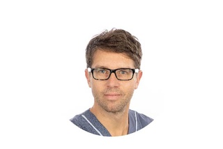 Person som bär glasögon och en blå skjorta, med kort brunt hår, mot en vit bakgrund.