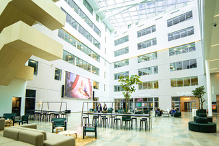 Modernt atrium med takfönster, sittplatser, bord, en stor skärm och krukträd. Ljus atmosfär med naturligt ljus.