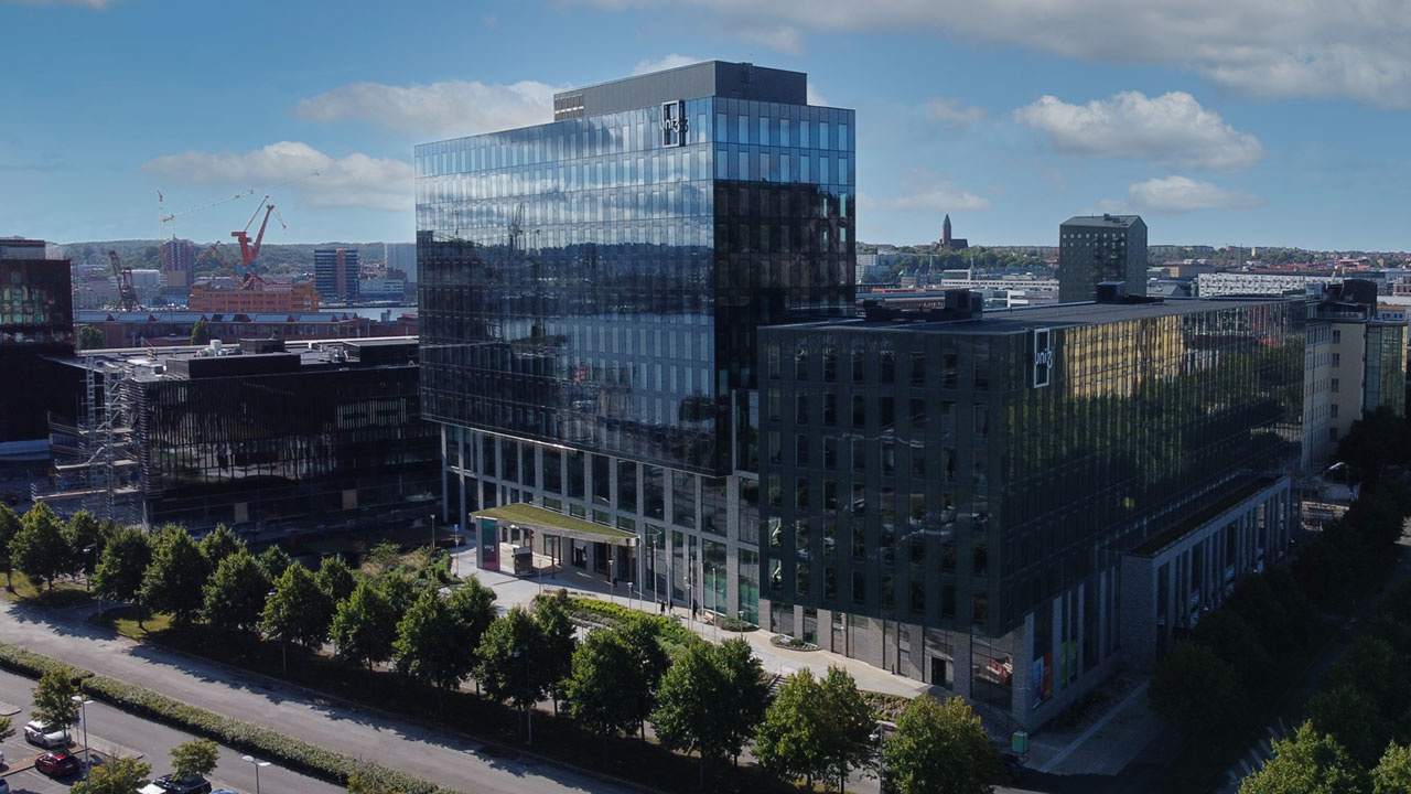 Kontorsbyggnad i glas som reflekterar himlen, omgiven av träd och andra stadsstrukturer under en delvis molnig himmel.