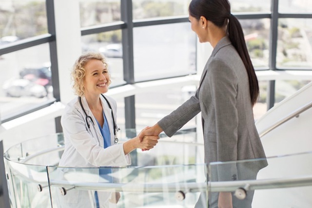 Doctor shaking hands with a woman in an office building, both smiling, with large windows in the background.