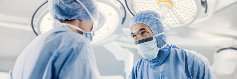 Zwei Chirurgen in blauer OP-Kleidung und Masken stehen in einem Operationssaal mit hellem Deckenlicht.