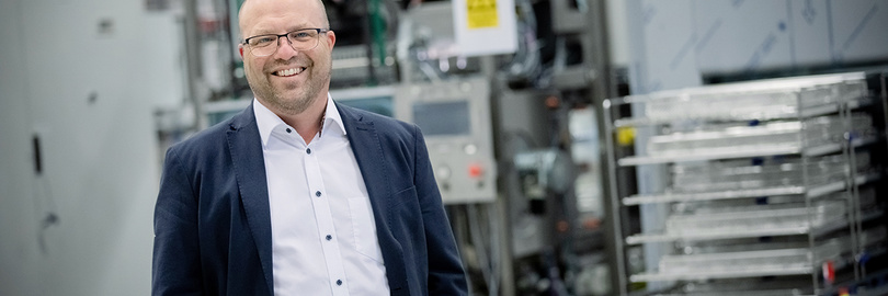 Lächelnder Mann in Anzug und Brille, der in einer Industrieanlage mit Maschinen im Hintergrund steht.