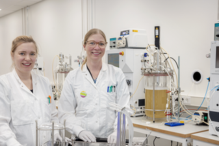 Twee vrouwen in witte jassen lachen in een laboratorium met grote bioreactor op een tafel.