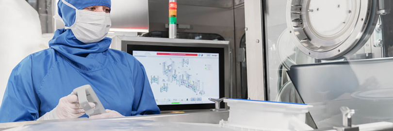 Persoon in blauwe beschermende kleding bedient machines in een steriele laboratoriumomgeving, met een monitor waarop diagrammen worden weergegeven.