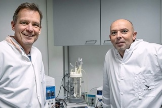 Twee mannen in laboratoriumjassen lachen naar de camera. Ze staan in een laboratorium met een bioreactor op de achtergrond.