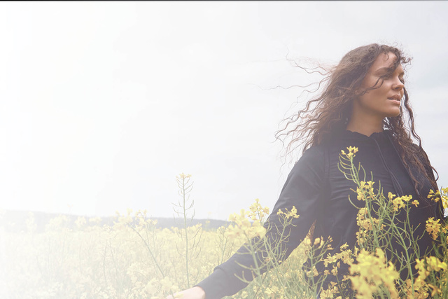 A woman with curly hair stands in a field of yellow flowers, wearing a black top, with cloudy sky in the background.