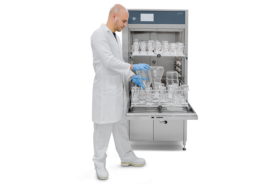 Person in lab coat and gloves loading glassware into a scientific dishwasher.