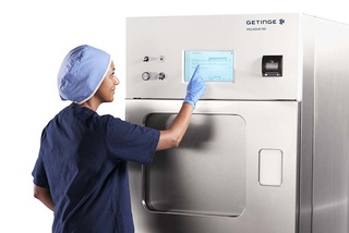 A person in scrubs and gloves operates a large medical sterilization machine with a touchscreen display.