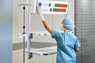 A medical professional adjusts equipment in a hospital room with an IV stand and control panel.