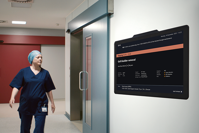 In an OR hallway is a nurse passing the digital door sign next to a OR door
