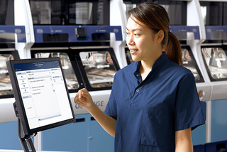 A woman in a blue uniform operates a touchscreen in a lab with machines in the background.
