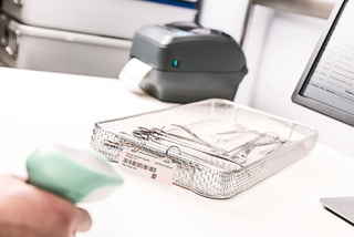 A person scans a barcode on a tray of medical instruments next to a monitor and a label printer.