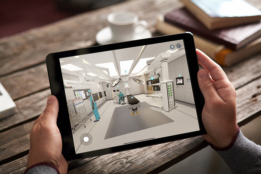 Person holding a tablet displaying a virtual surgery room simulation on a wooden table.