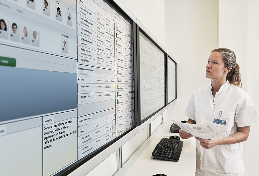 A healthcare professional in a white coat reviews data on large wall-mounted screens in a medical facility.
