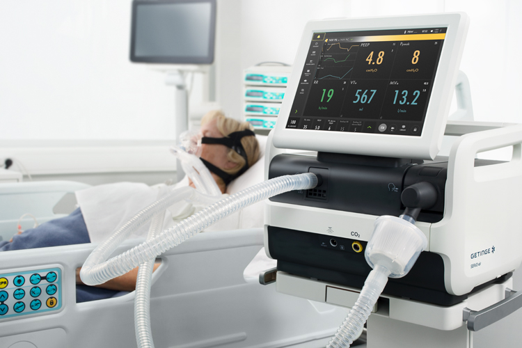 Adult patient in hospital bed wearing non-invasive mask and Getinge Servo-air co2 mechanical ventilator next to the bed