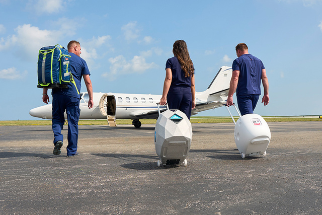 Three medical professionals in blue scrubs walk toward a jet on a tarmac, carrying a large backpack and two advanced Paragonix Organ Preservation devices