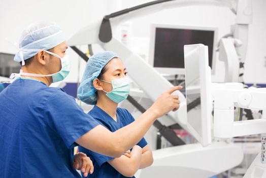Two medical professionals in surgical attire discuss a screen in a hospital setting.