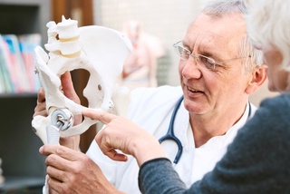 Doctor explaining hip joint model to a patient in an office filled with books.