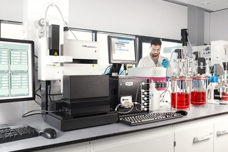 Scientist in a lab working with computers and equipment, with red liquid-filled glassware in the foreground.