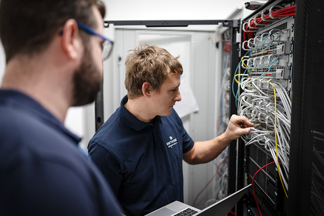 Two Getinge technicians in a server room work with network cables; one holds a laptop while adjusting cables on a server rack, the other observes.