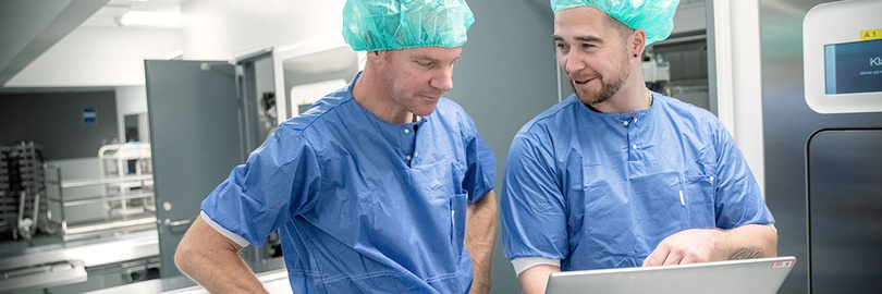 Two surgeons in surgical scrubs focused on a laptop screen in a clinical environment.
