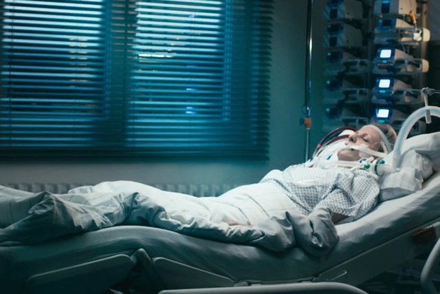 The image shows a patient lying in an ICU bed in a dimly lit hospital room, connected to life-support equipment.